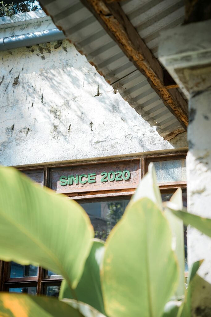 Sunlit rustic building with tropical leaves and 'Since 2020' text on the wall.